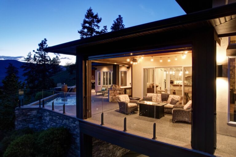 Covered outdoor living area at dusk with a firepit table and wicker seating, opening to a lit swimming pool and hillside view.