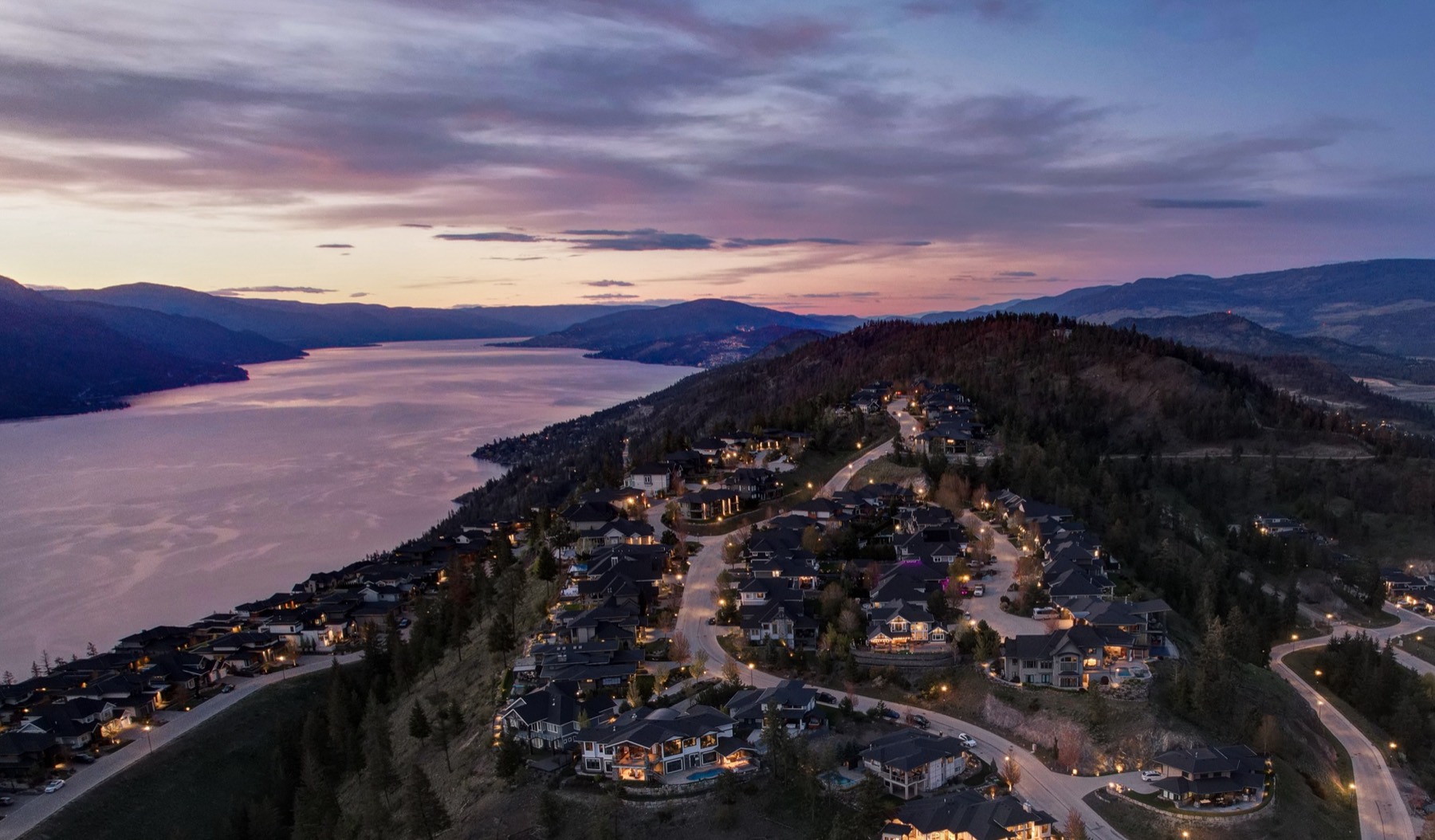 Aerial view at twilight of a hillside neighbourhood above Okanagan Lake, with lit homes following curved streets down the slope and mountains beyond.