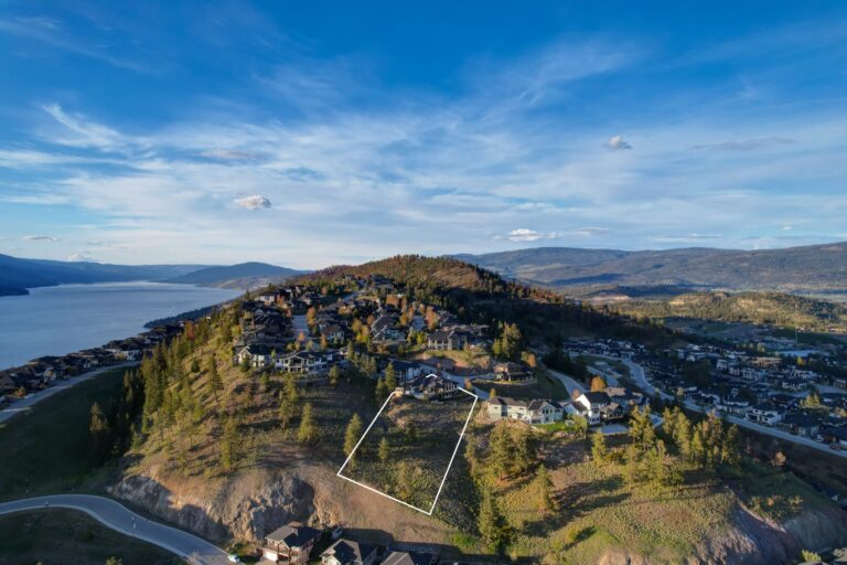 Aerial photograph of a hillside with a vacant building lot outlined in white, surrounded by large homes, with Okanagan Lake in the distance.
