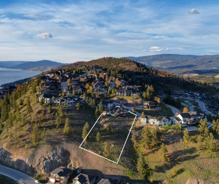 Aerial view of large homes spread along a forested hillside above Okanagan Lake, with a vacant lot outlined in white among them.