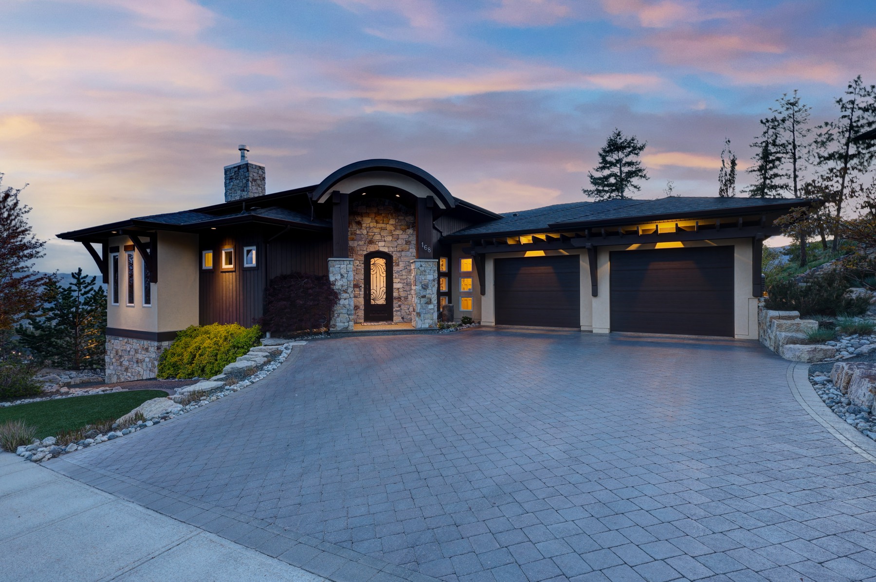 Front exterior of a single-storey home at twilight, with a stone entry archway, timber beams, a double garage and a paved driveway.