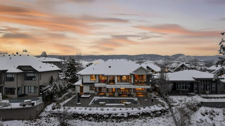 Rear elevation of a large home at twilight in winter, lit from inside, with a snow-edged swimming pool and patio and hills behind.