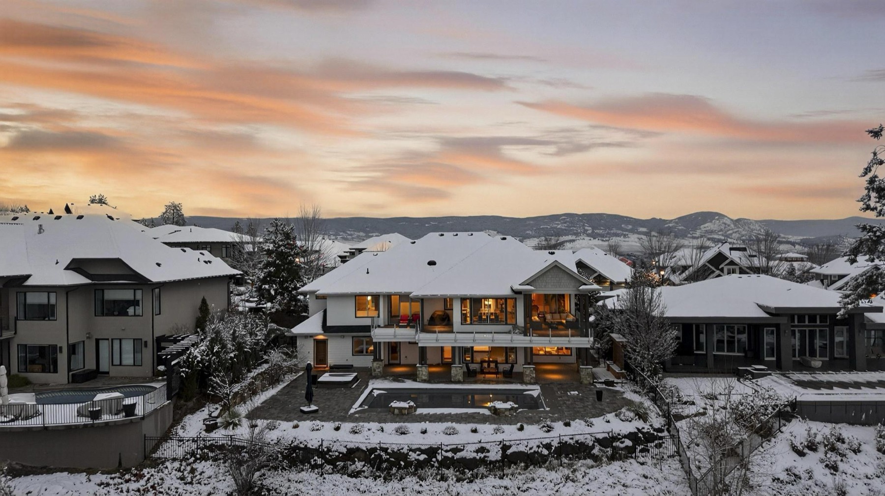 Rear elevation of a large home at twilight in winter, lit from inside, with a snow-edged swimming pool and patio and hills behind.