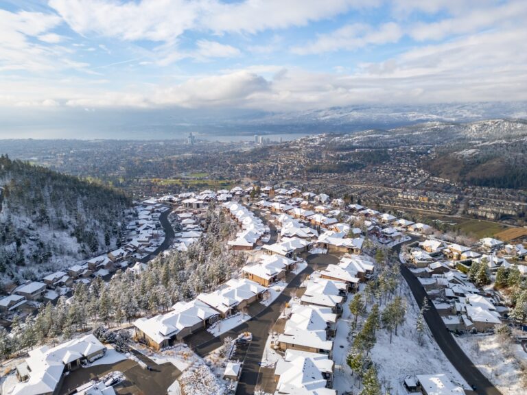 Aerial view of a hillside neighbourhood under fresh snow, with snow-covered roofs and pines, and the city and Okanagan Lake in the valley below.