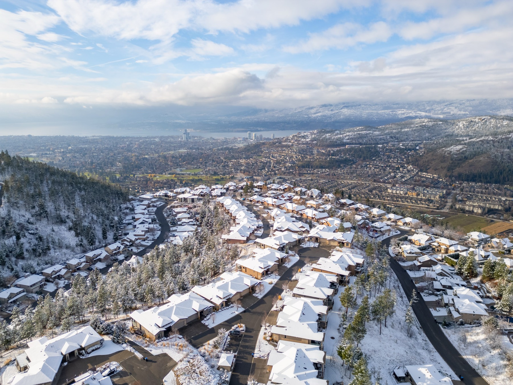 Aerial view of a hillside neighbourhood under fresh snow, with snow-covered roofs and pines, and the city and Okanagan Lake in the valley below.