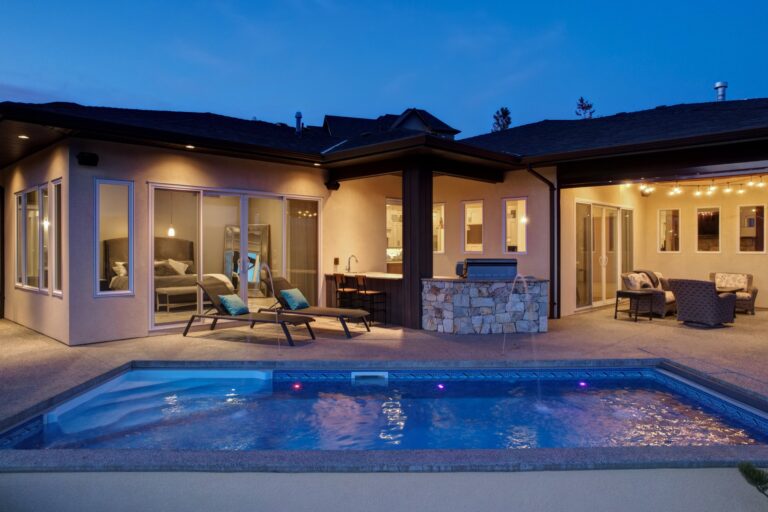 Rear of a single-storey home at dusk, lit from inside, with a swimming pool, loungers and a covered outdoor kitchen.