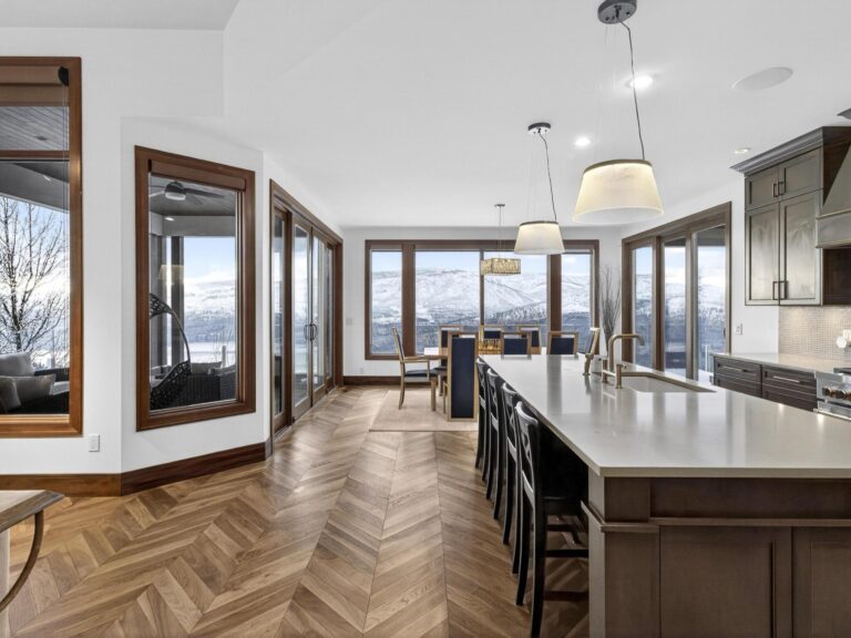 Open kitchen and dining area with a long quartz island, herringbone wood floors and floor-to-ceiling windows looking out to snow-covered hills.