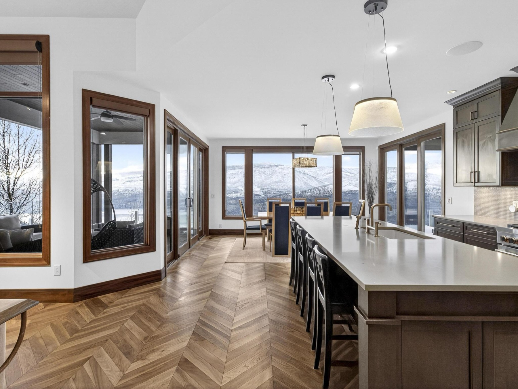 Open kitchen and dining area with a long quartz island, herringbone wood floors and floor-to-ceiling windows looking out to snow-covered hills.