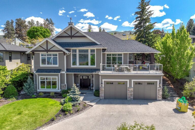 Front exterior of a two-storey Kelowna home with grey siding, timber-accented gables, a covered upper deck and a three-car garage.