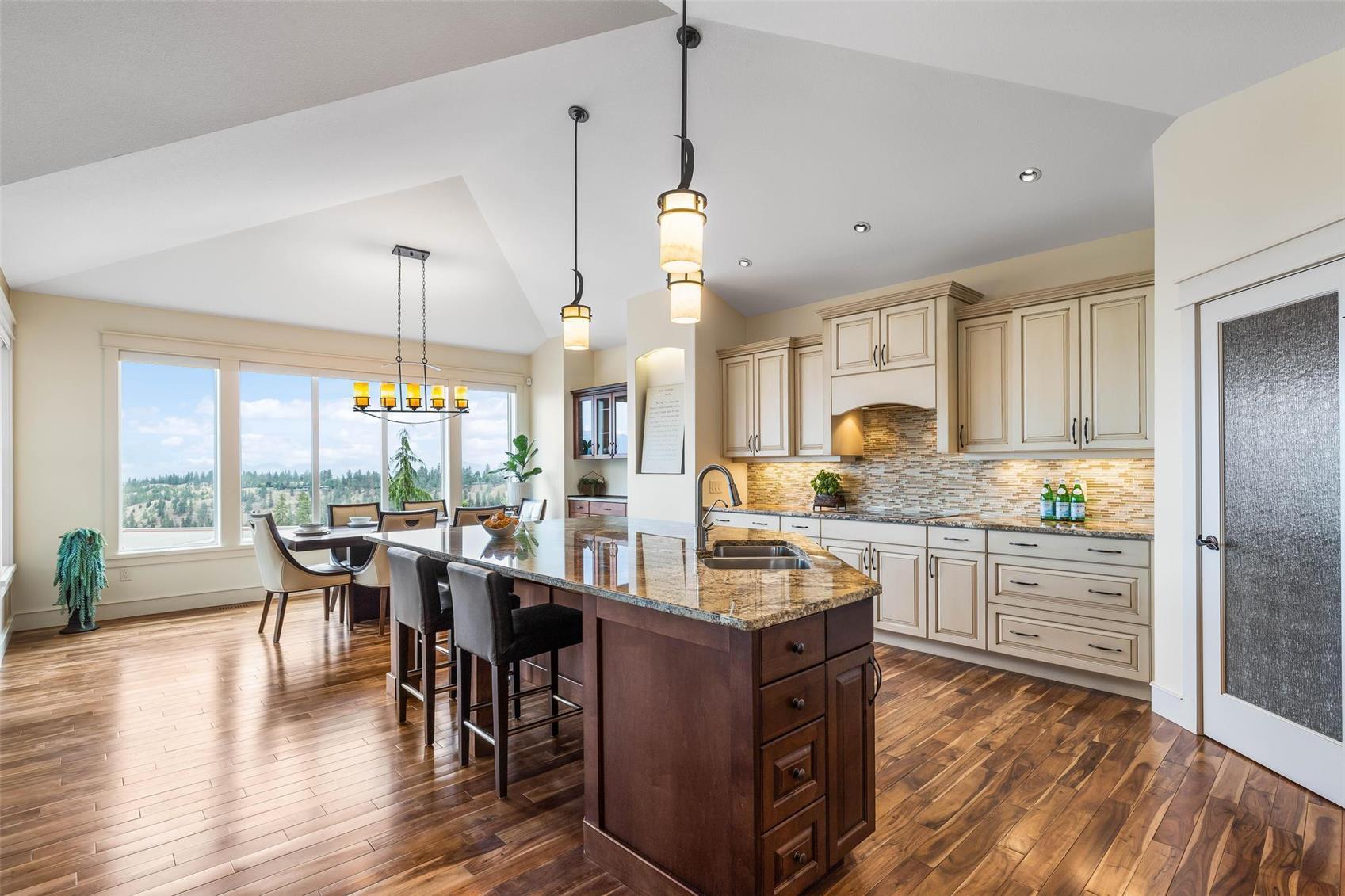 Open-plan kitchen and dining area with a granite island, dark hardwood floors and vaulted ceiling, with dry Okanagan hillside and pines visible through the windows.