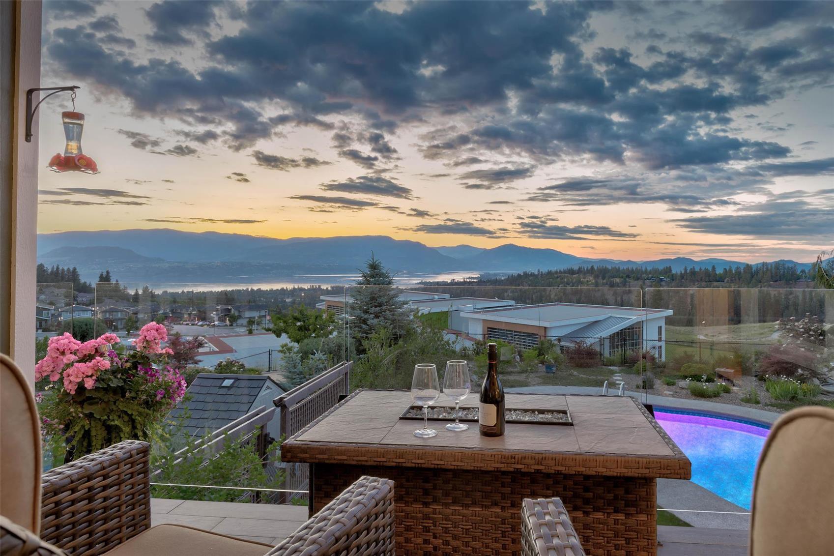 Covered patio at twilight looking out over Okanagan Lake and the surrounding hills, with a lit swimming pool below and a table set with wine glasses.