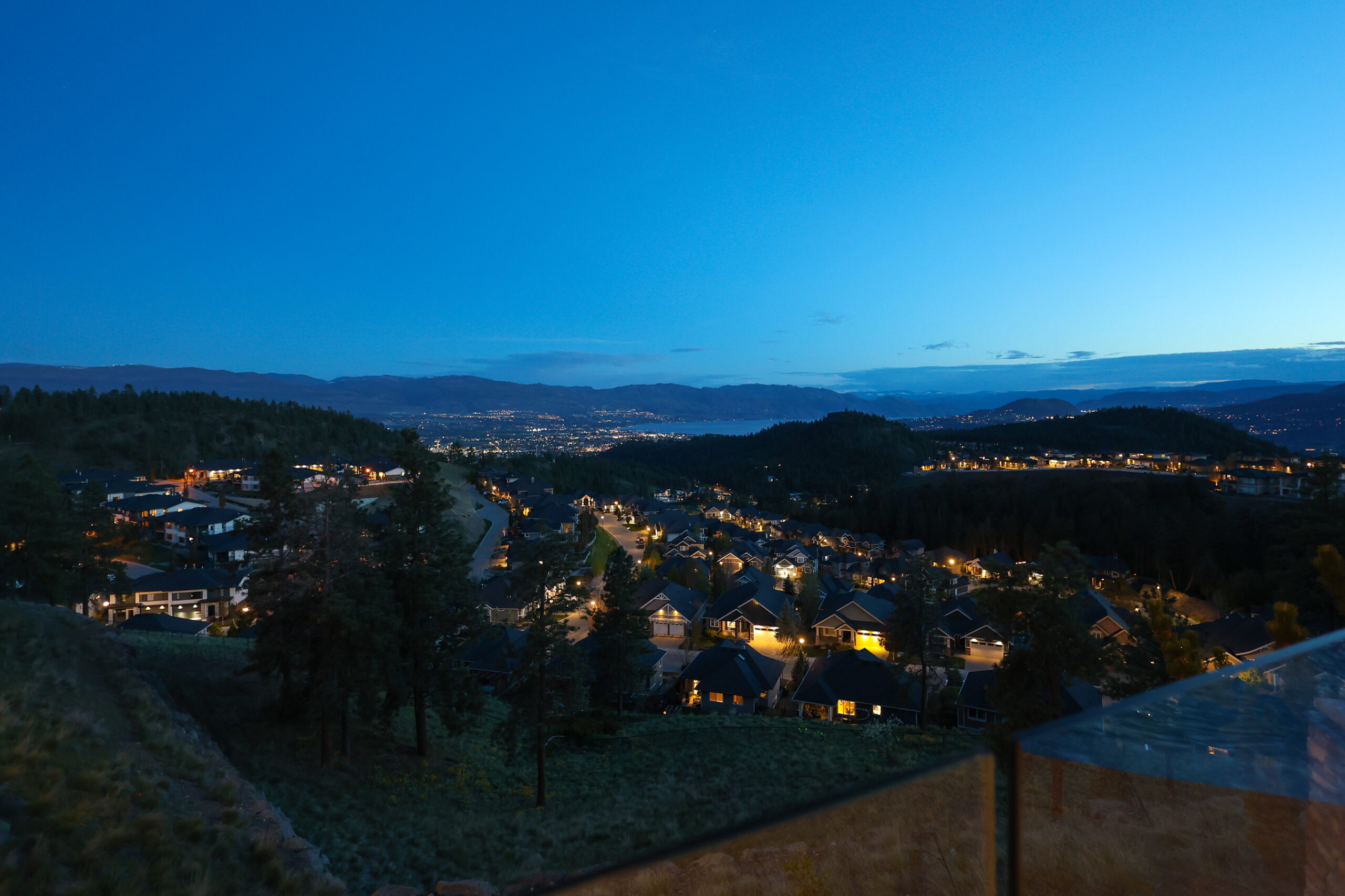 Upper Mission hillside homes at dusk, looking down over rooftops toward Okanagan Lake and the lights of Kelowna.