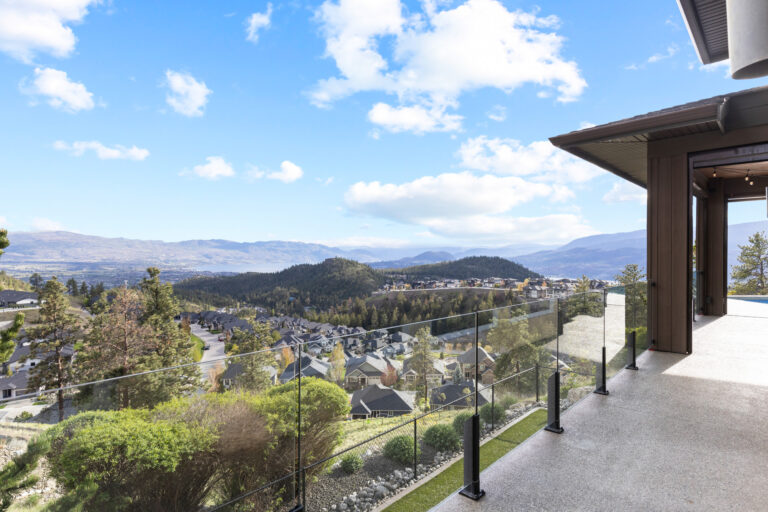 Covered terrace with a glass railing looking out over a hillside neighbourhood of homes toward Okanagan Lake
