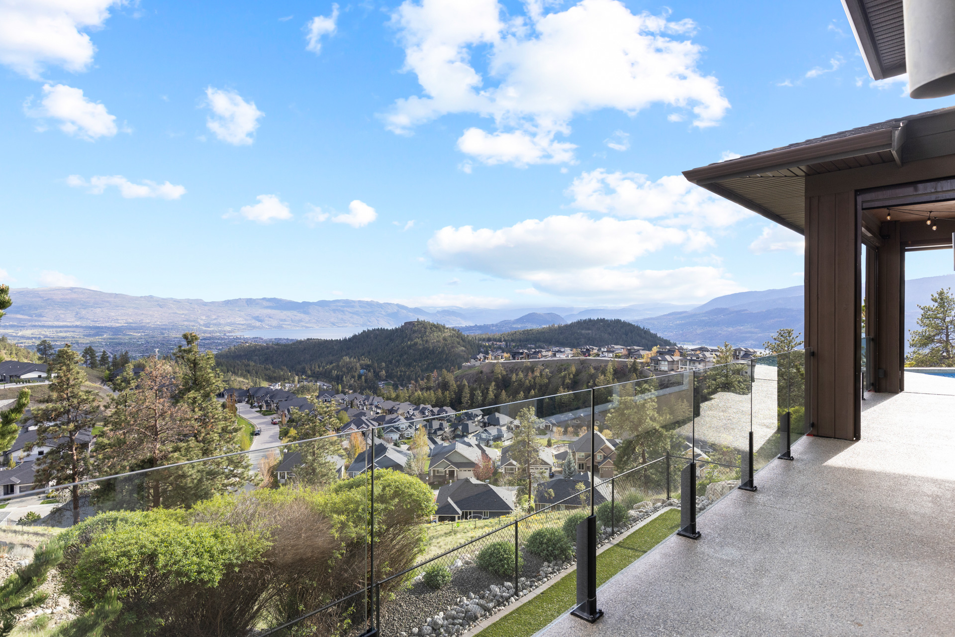 Covered terrace with a glass railing looking out over a hillside neighbourhood of homes toward Okanagan Lake