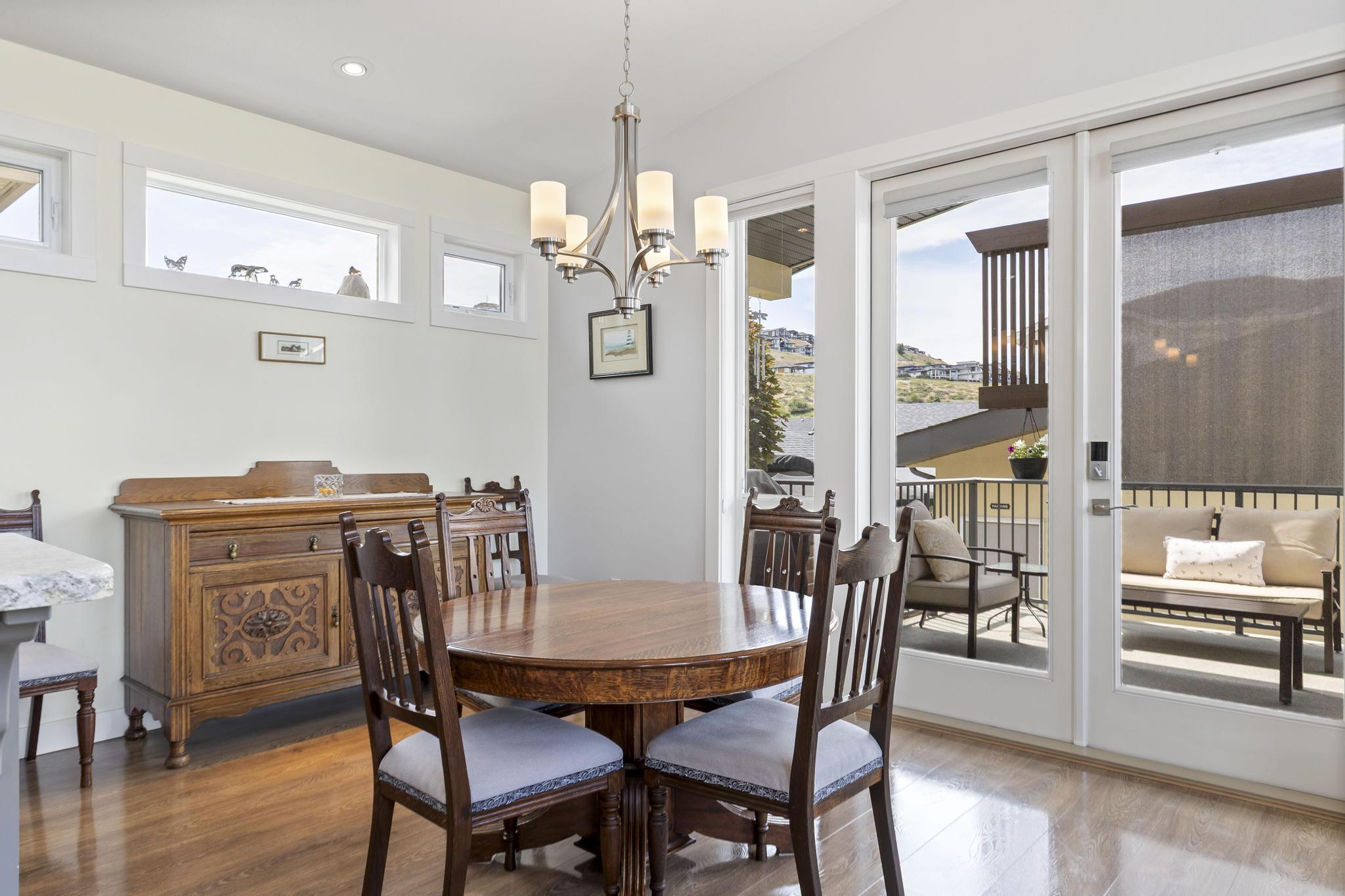 Dining area of a custom-built Upper Mission rancher, with french doors open to a covered deck and the dry Okanagan hillside and neighbouring rooflines beyond.